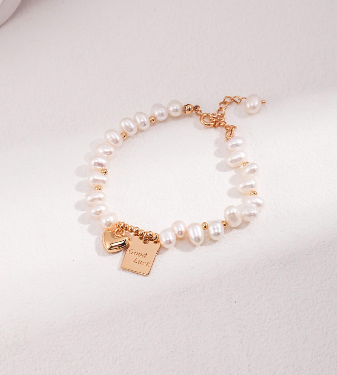 Natural pearl bracelet with a gold-tone heart charm and engraved Good Luck tag on a pale background.