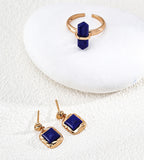 A pair of 18K gold-plated drop earrings with square-cut dark blue lapis lazuli stones, displayed on a textured white surface next to a matching gold ring with a vertical dark lapis lazuli stone, both resting on a smooth white stone.