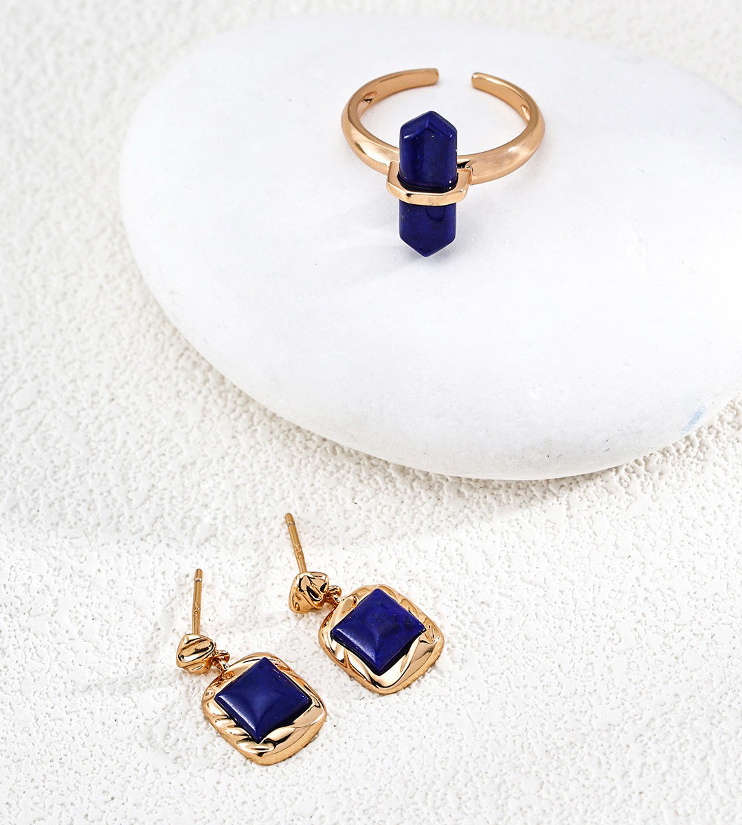 A pair of 18K gold-plated drop earrings with square-cut dark blue lapis lazuli stones, displayed on a textured white surface next to a matching gold ring with a vertical dark lapis lazuli stone, both resting on a smooth white stone.
