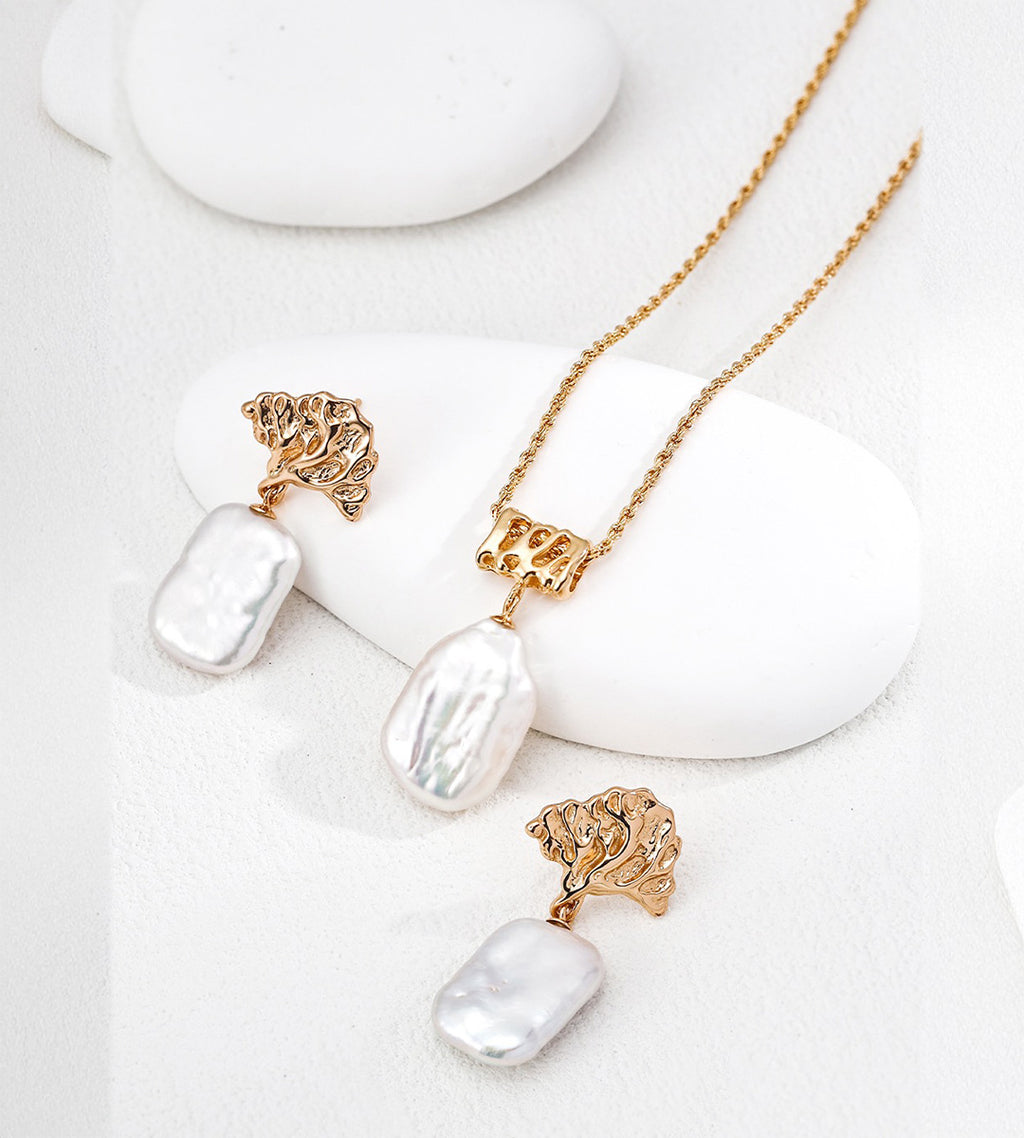 Baroque pearl rope chain necklace displayed beside pearl drop earrings with sculptural gold-tone tops on white display blocks.