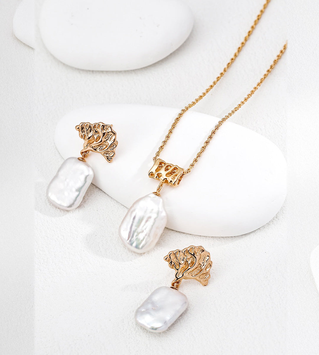 Baroque pearl rope chain necklace displayed beside pearl drop earrings with sculptural gold-tone tops on white display blocks.