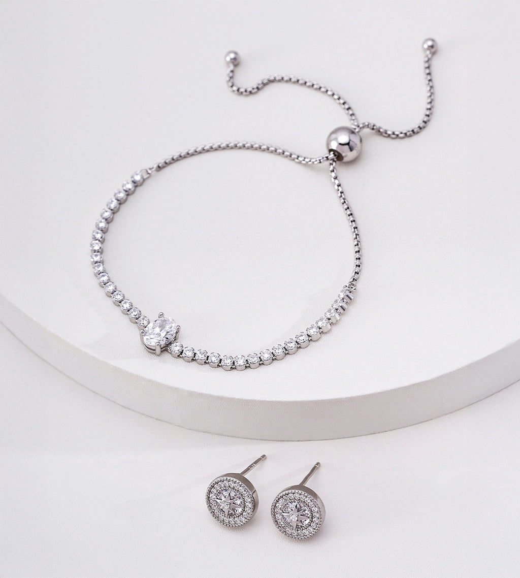 Silver-tone oval cubic zirconia tennis bracelet on a white display, with a sliding ball closure and circular stud earrings below.


