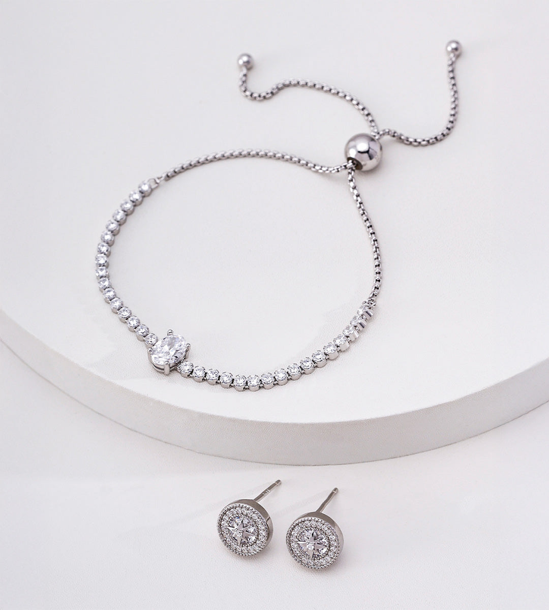 Silver-tone oval cubic zirconia tennis bracelet on a white display, with a sliding ball closure and circular stud earrings below.

