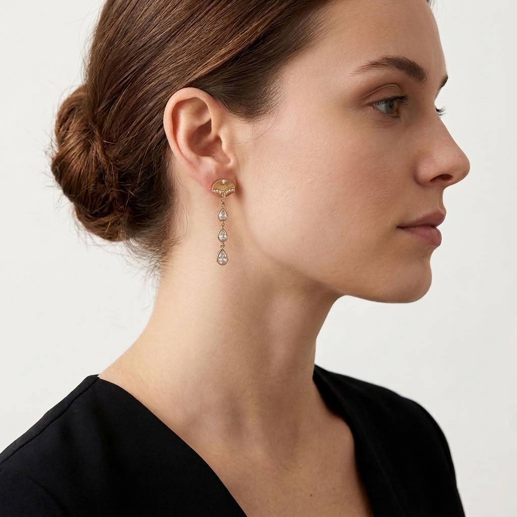 A close-up side profile of a woman wearing a delicate gold dangle earring with three clear teardrop-shaped gemstones, set against a clean, light off-white background.