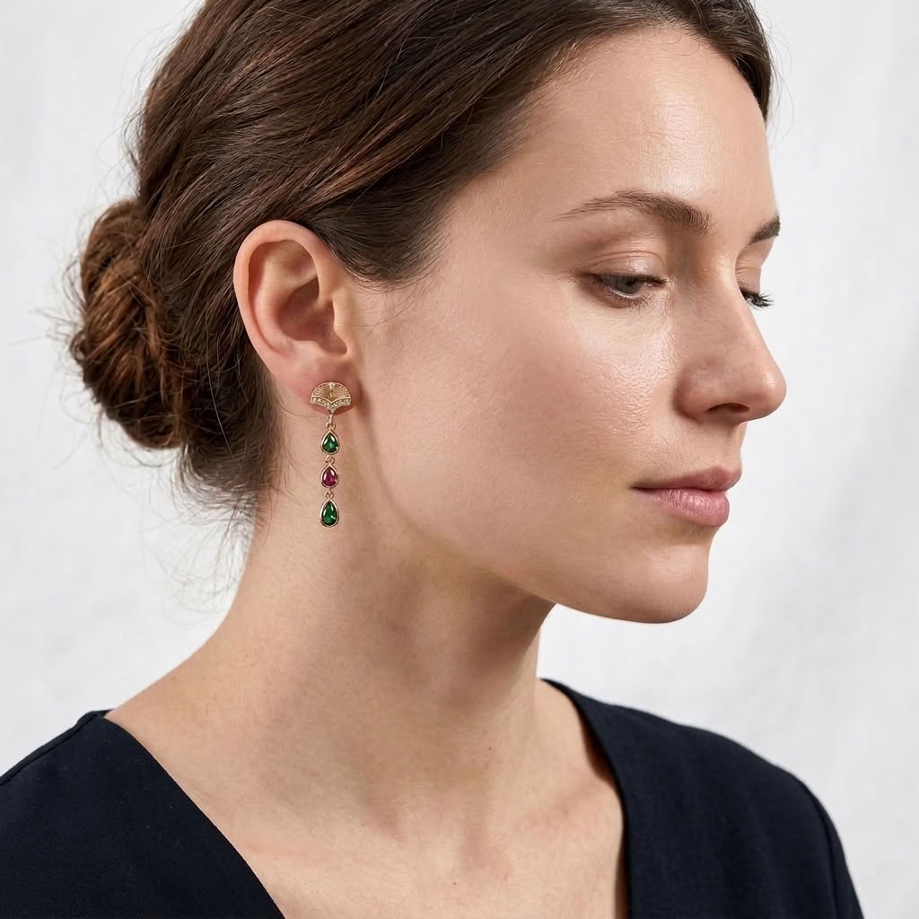 A woman in profile wearing a delicate gold dangle earring with small green and red gemstones, shown against a soft light gray background.