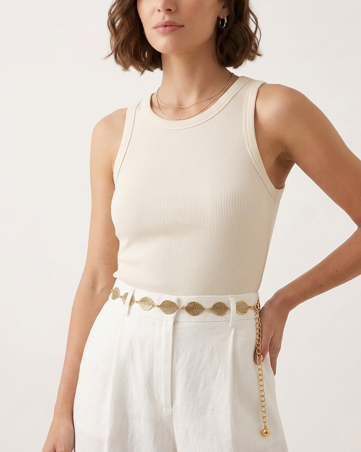 A woman wearing a ribbed cream sleeveless top and white trousers, accessorized with a Bohemian gold leaf waist belt at the waist | BUCKLE Collective