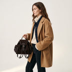 A woman wearing a camel coat, cream turtleneck, and dark denim holds a dark brown suede backpack by BUCKLE Collective