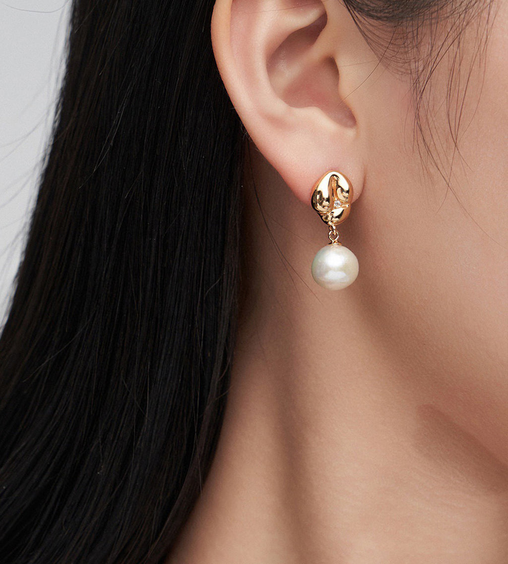 A close-up of a woman's ear wearing an 18K gold-plated drop earring with a dangling white pearl.