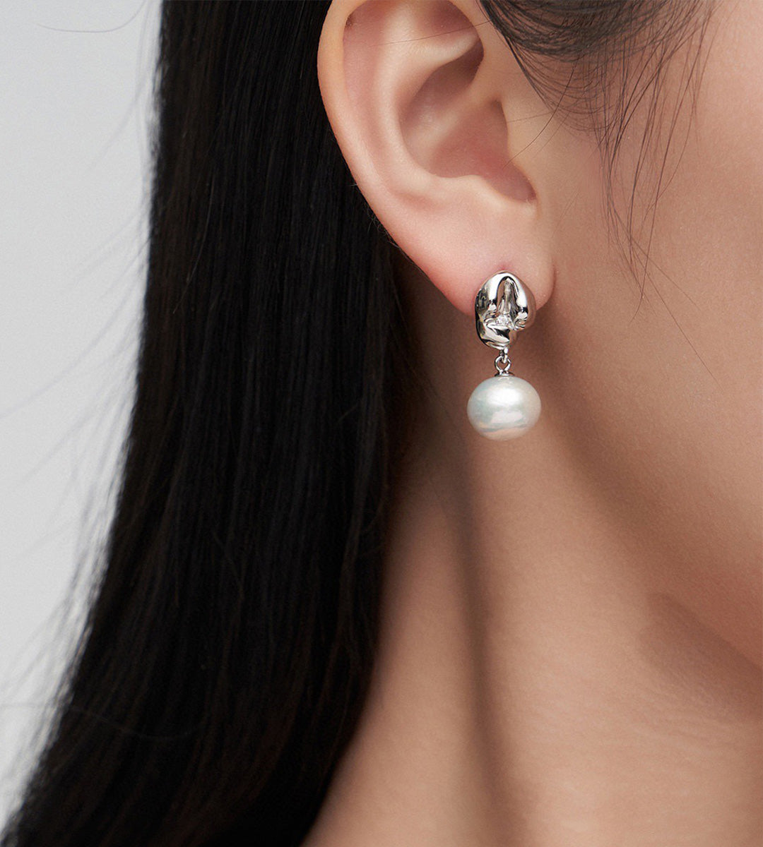 A close-up of a woman's ear wearing an 18K white-gold-plated drop earring with a dangling white pearl.