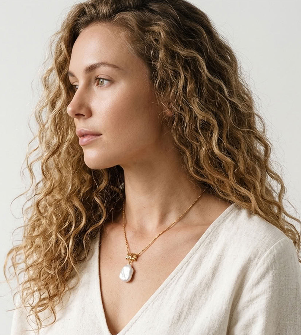 Model wearing a baroque pearl pendant necklace with a gold-tone rope chain and sculptural connector, styled with a white V-neck blouse.