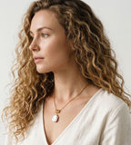 Model wearing a baroque pearl pendant necklace with a gold-tone rope chain and sculptural connector, styled with a white V-neck blouse.