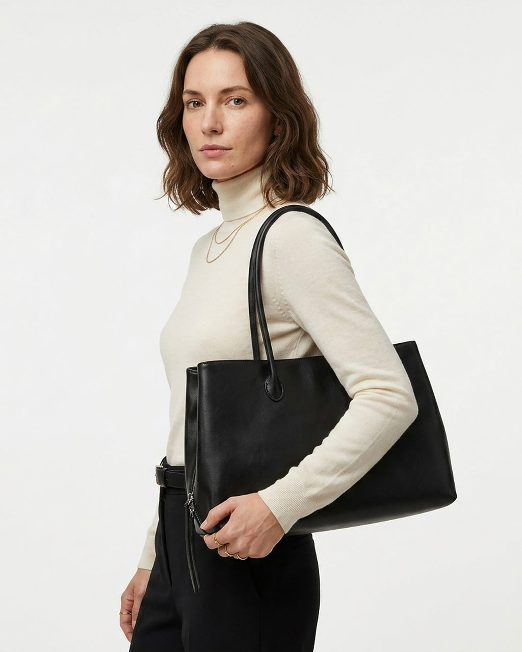 A woman wearing a cream turtleneck sweater and black trousers carries a large black leather commuter tote bag over her shoulder. BUCKLE Collective