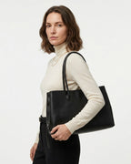 A woman wearing a cream turtleneck sweater and black trousers carries a large black leather commuter tote bag over her shoulder. BUCKLE Collective