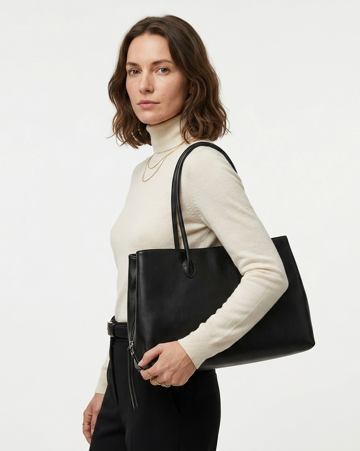 A woman wearing a cream turtleneck sweater and black trousers carries a large black leather commuter tote bag over her shoulder. BUCKLE Collective