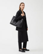 A model in a black blazer, long-sleeve top, and trousers carries a large black woven leather tote bag