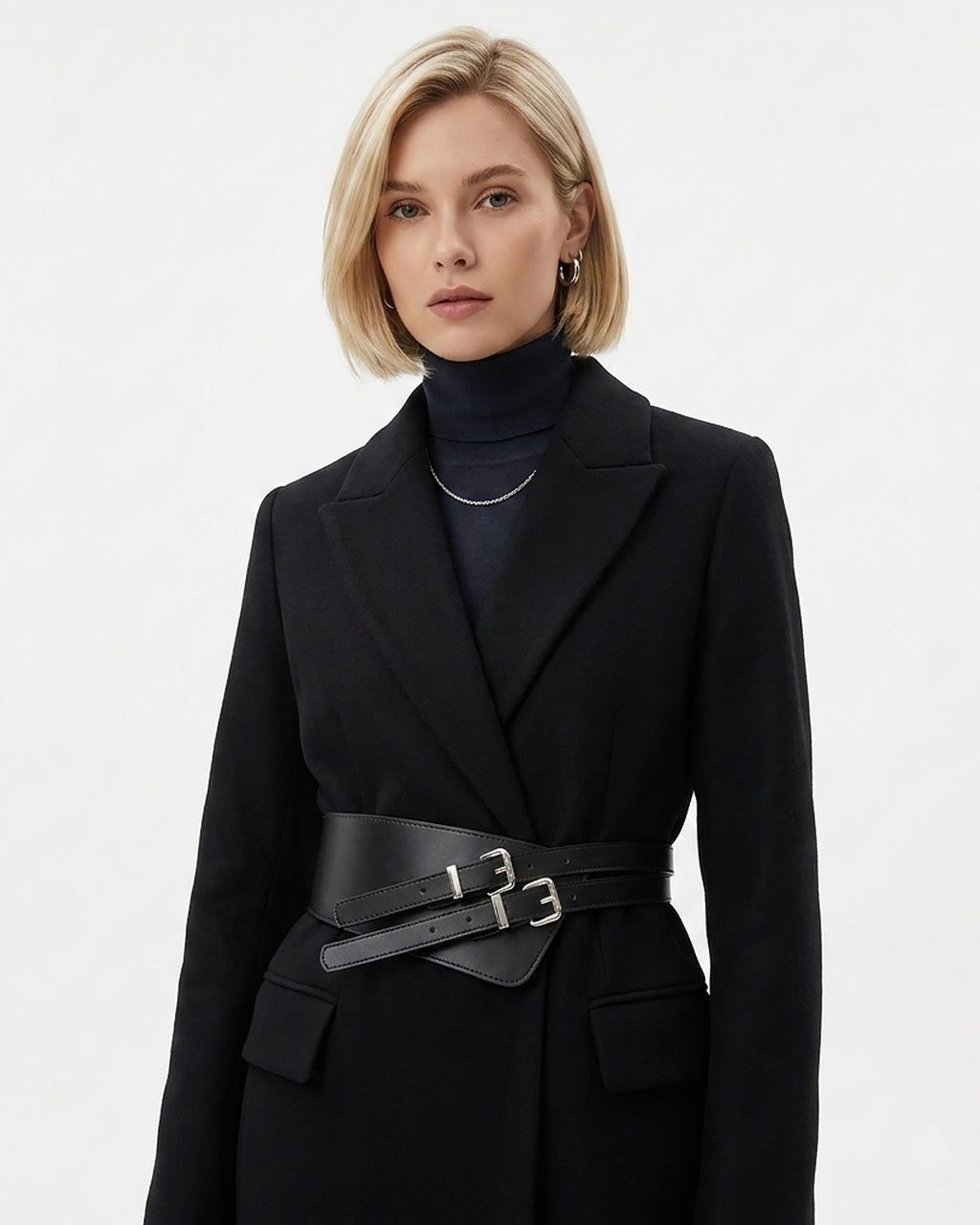 A blonde model wearing a black double-breasted coat with a wide genuine leather black belt featuring double silver buckles, styled over a dark navy turtleneck sweater | BUCKLE Collective