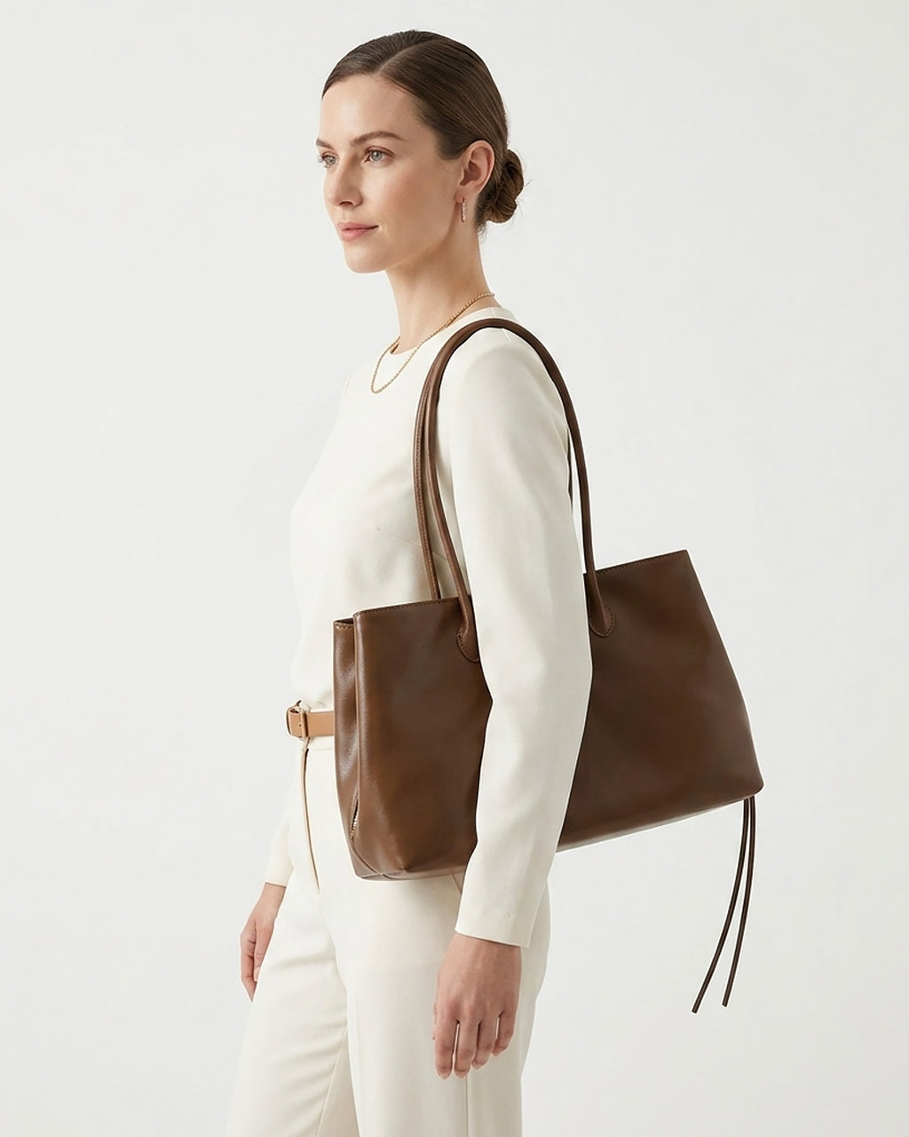 A woman in a cream-colored outfit is shown from the side, carrying a large brown leather tote bag over her shoulder. BUCKLE Collective