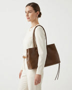 A woman in a cream-colored outfit is shown from the side, carrying a large brown leather tote bag over her shoulder. BUCKLE Collective