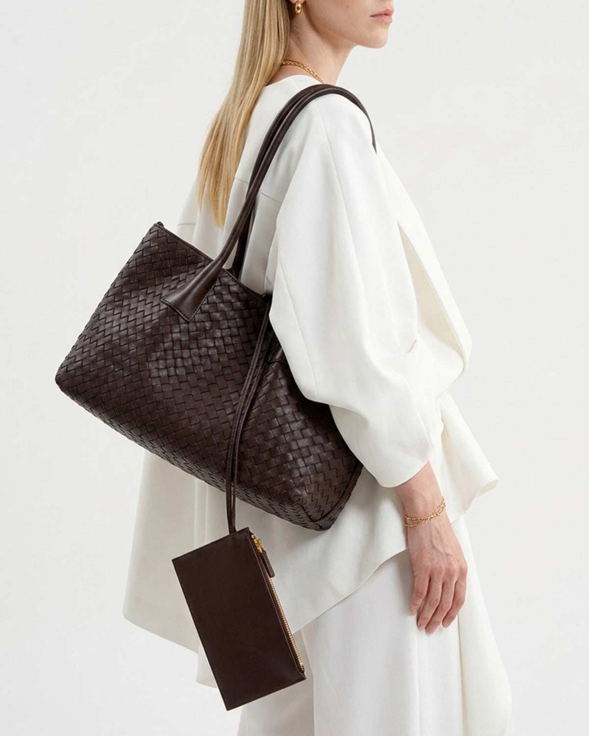 A woman in a white draped outfit carries a dark brown woven leather tote bag with a matching slim wallet or pouch hanging from the strap.