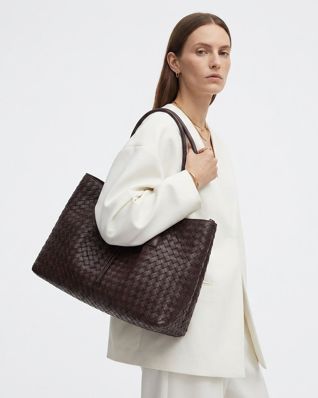 A woman wearing a white blazer carries a large dark brown woven leather tote bag over her shoulder