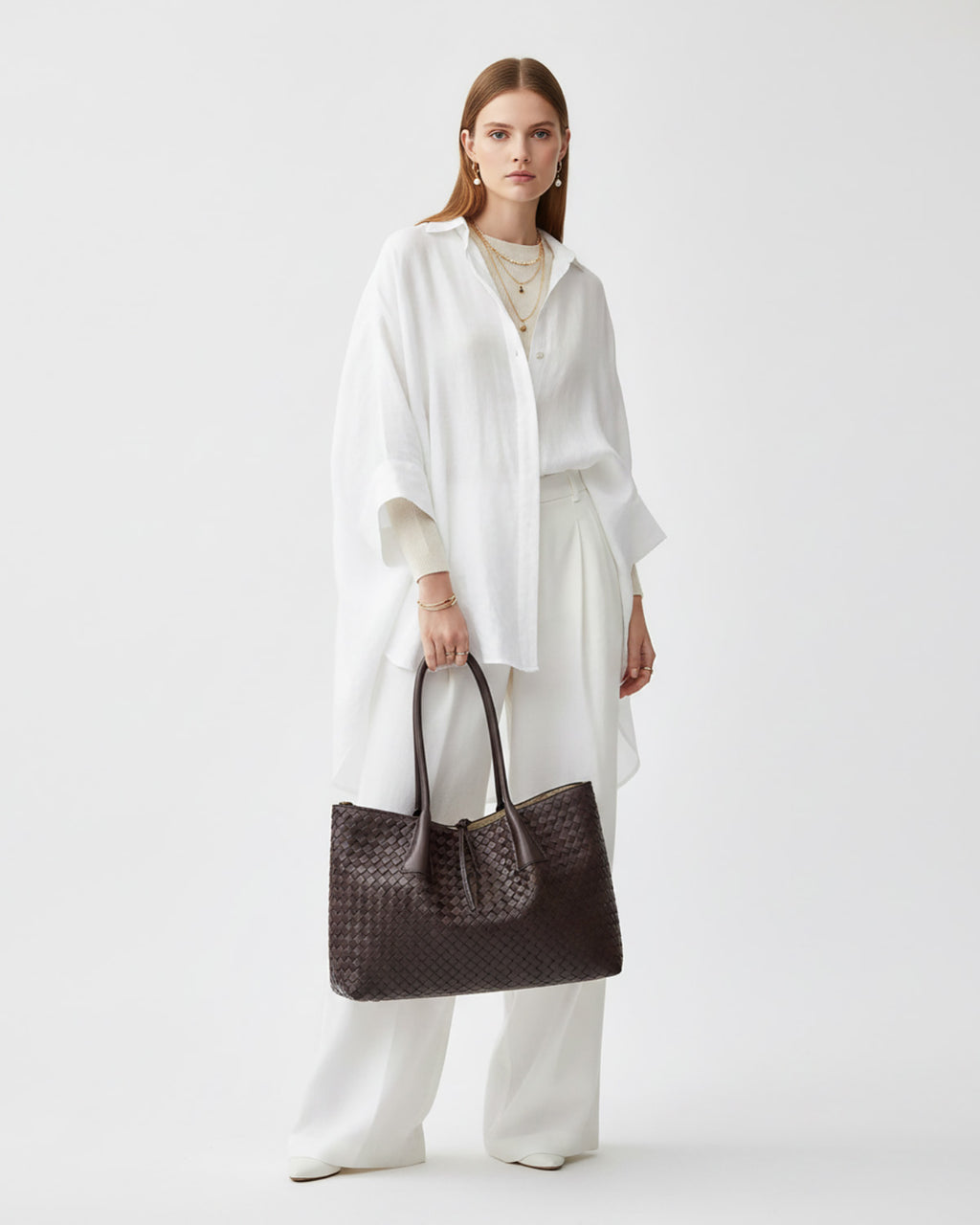 A model stands against a light gray studio background wearing a white draped shirt, white wide-leg pants, and holding a large dark brown woven tote bag.