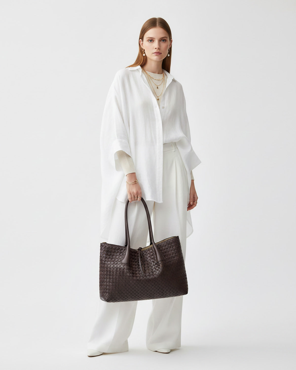 A model stands against a light gray studio background wearing a white draped shirt, white wide-leg pants, and holding a large dark brown woven tote bag.