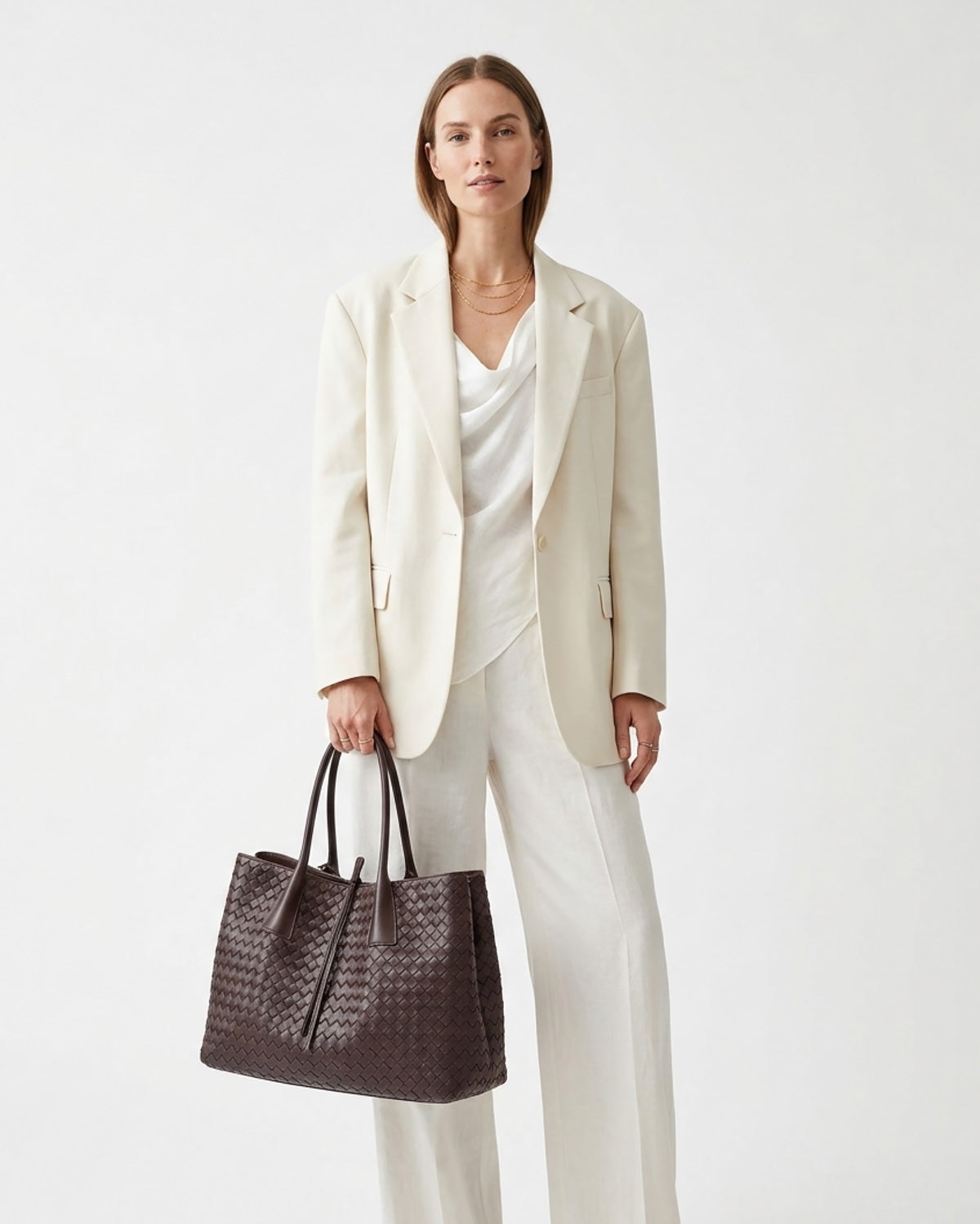 A woman stands against a plain light background wearing an off-white blazer, white top, and white wide-leg trousers, while holding a dark brown woven tote bag.