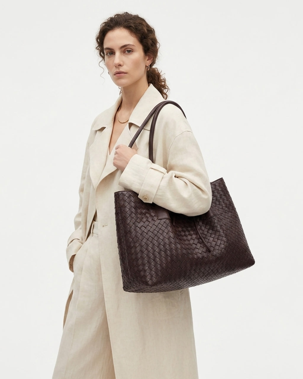 A woman wearing a white blazer carries a large dark brown woven leather tote bag over her shoulder