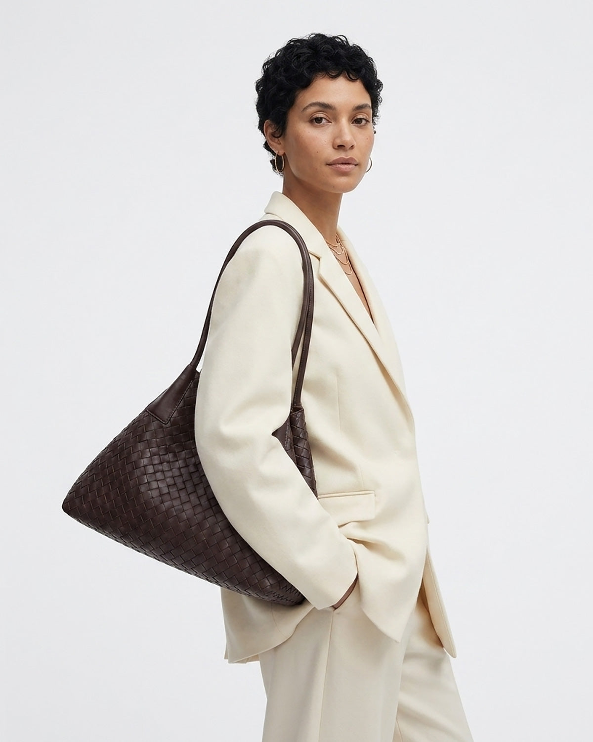 A model wearing a cream-colored blazer and trousers carries a large, dark brown woven leather tote bag