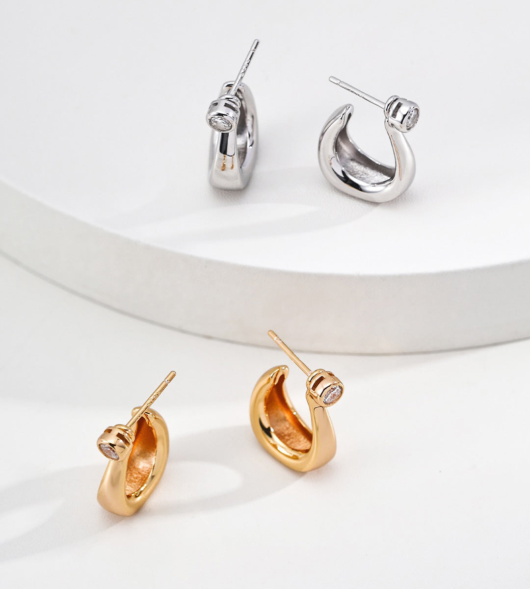 Two pairs of hoop-style earrings are displayed on a clean white surface. Each earring has a small, clear cubic zirconia stone set near the hinge.