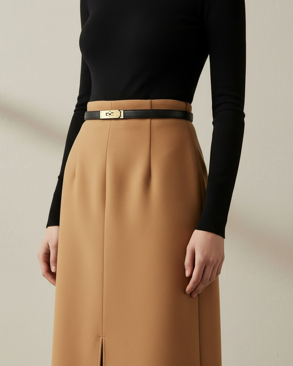A model wearing a black long-sleeve top and a high-waisted camel brown skirt with a thin Black Classic Saffiano Leather Belt with a gold turn-lock buckle