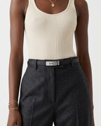 A close-up of a model wearing a cream ribbed tank top, dark charcoal gray wool trousers, and a black saffiano leather belt with a silver-tone turn-lock buckle