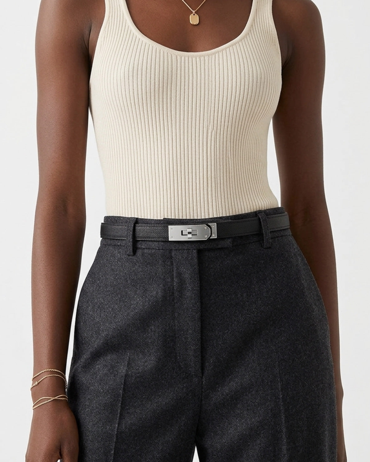 A close-up of a model wearing a cream ribbed tank top, dark charcoal gray wool trousers, and a black saffiano leather belt with a silver-tone turn-lock buckle