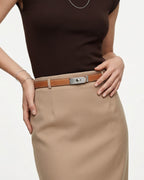 A close-up of a model wearing a tan slim saffiano leather belt with a silver-tone turn-lock buckle and contrast stitching, styled with a beige pencil skirt and a dark brown top against a light gray background.