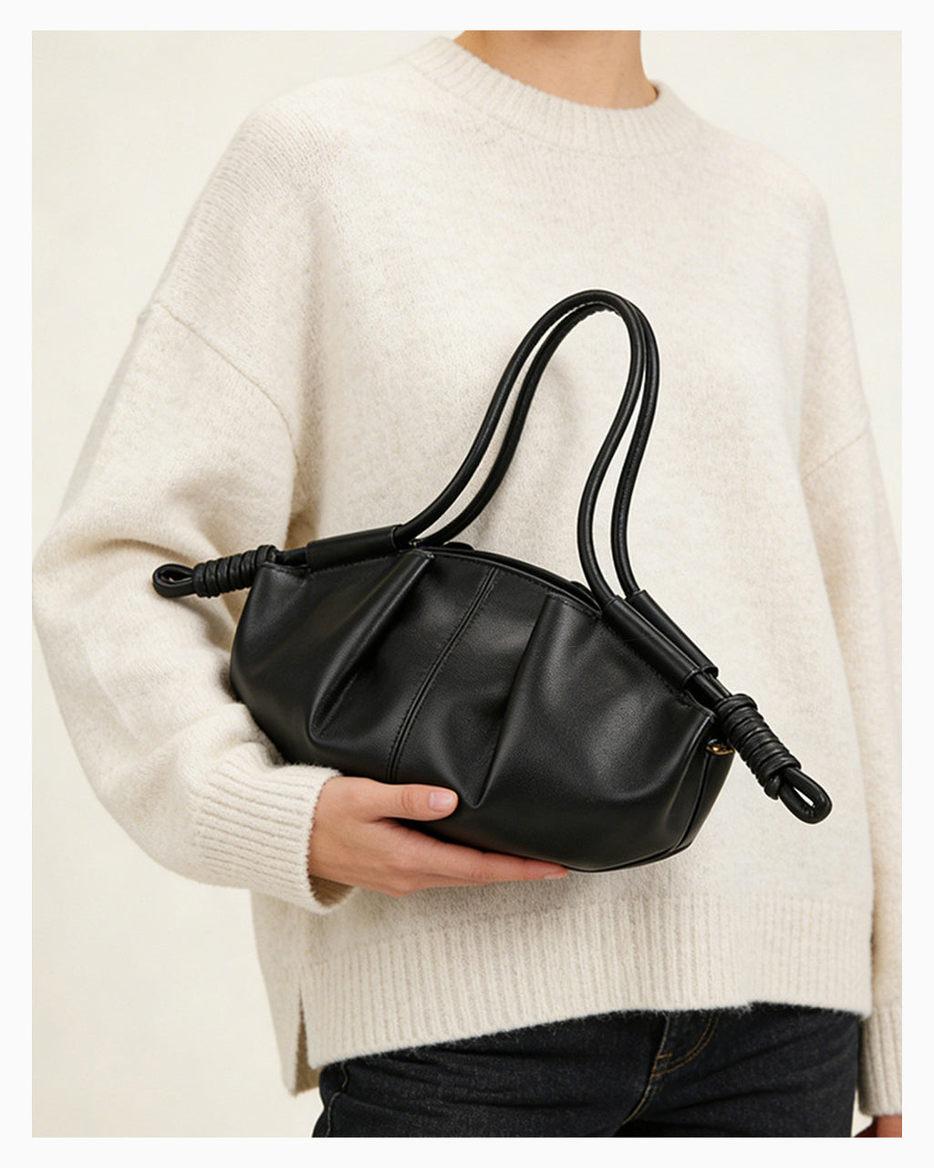 Woman in stylish knitwear and black jeans holds a black pleated dumpling leather handbag with a knotted strap