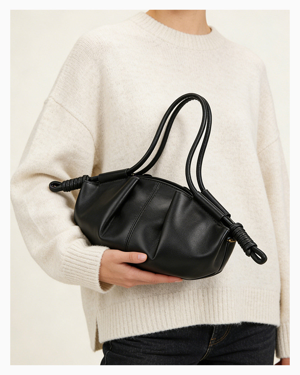 Woman in stylish knitwear and black jeans holds a black pleated dumpling leather handbag with a knotted strap