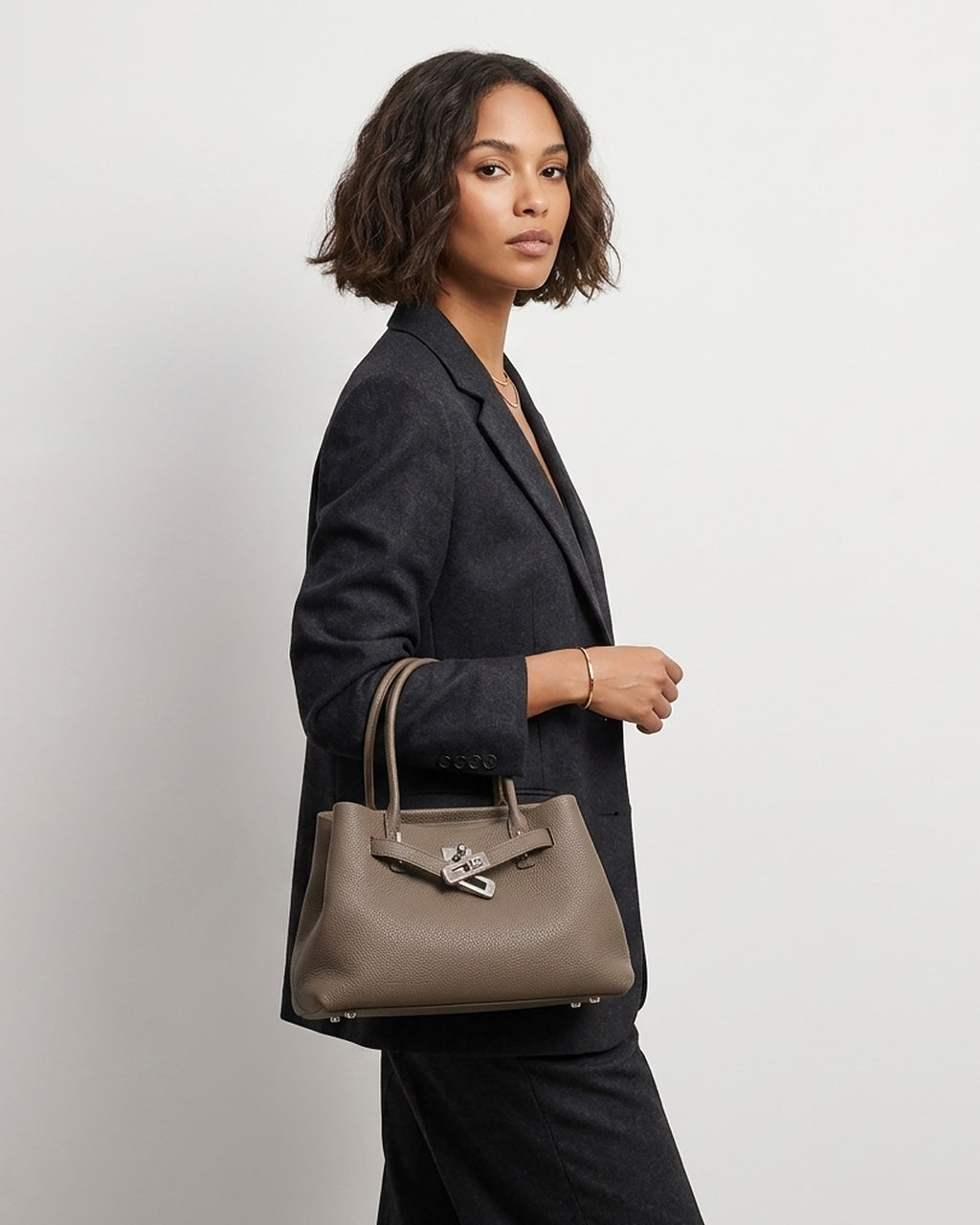 A woman in a dark gray blazer and black trousers carries an Elephant Gray Commuter Genuine Togo Leather Satchel Bag with Turn-Lock | BUCKLE Collective