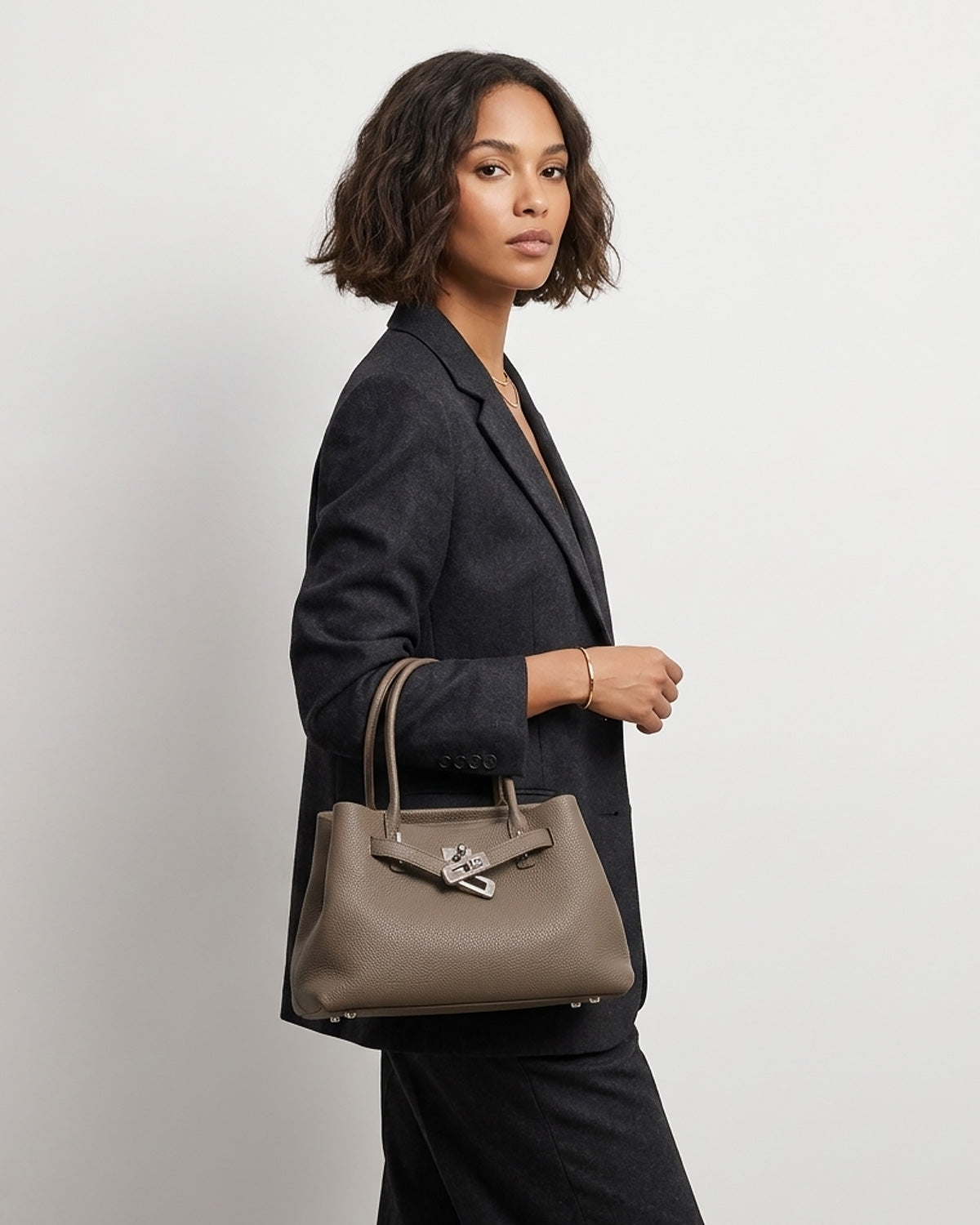 A woman in a dark gray blazer and black trousers carries an Elephant Gray Commuter Genuine Togo Leather Satchel Bag with Turn-Lock | BUCKLE Collective