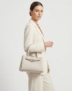 A woman in a cream-colored blazer and trousers carries an Off-White Commuter Genuine Togo Leather Satchel Bag with Turn-Lock | BUCKLE Collective