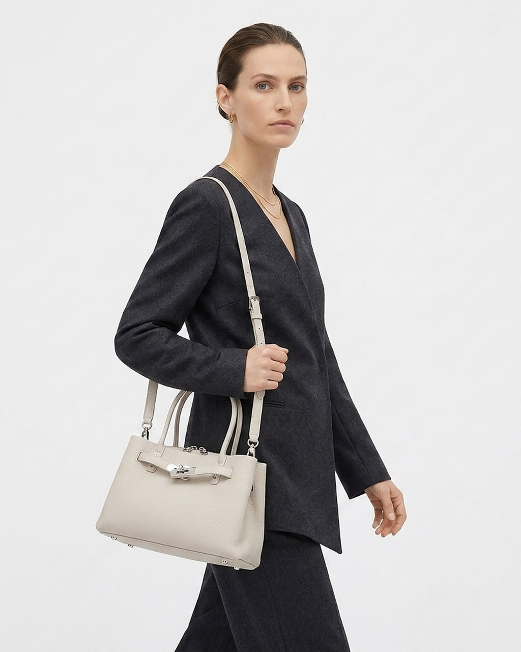 A model wearing a dark charcoal gray blazer and black trousers carries an Off-White Commuter Genuine Togo Leather Satchel Bag with Turn-Lock & Detachable Strap | BUCKLE Collective