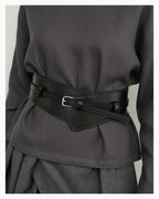 A woman in an elegant dark-gray outfit wearing a 2‑in‑1 transformable black leather corset belt with a gold buckle. The belt transforms from a statement, wide design into a slim classic design