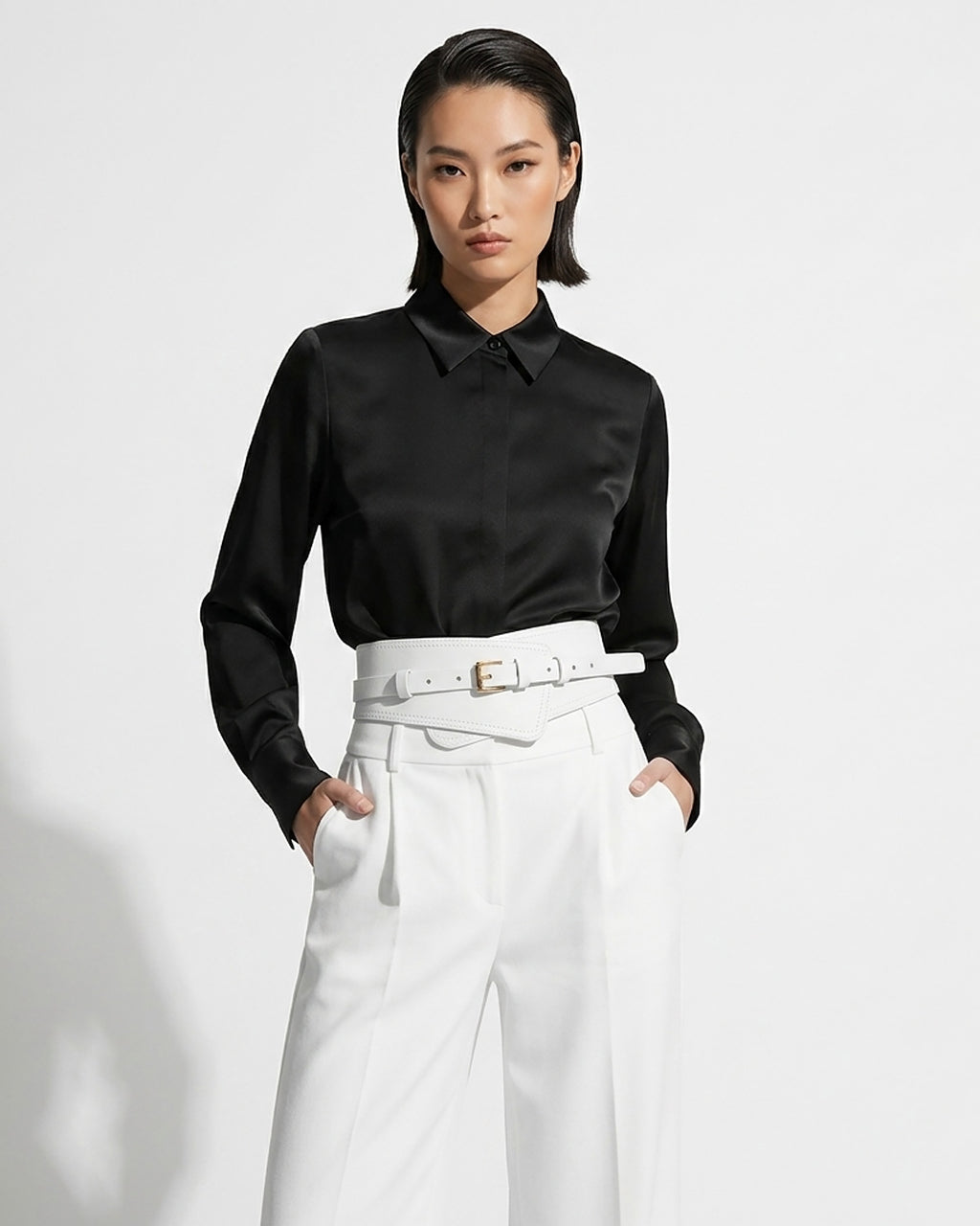A model wearing a black satin long-sleeve button-down shirt tucked into high-waisted white trousers with a matching 2‑in‑1 White Women’s Leather Wide Transformable Statement Belt with Gold Buckle | BUCKLE Collective