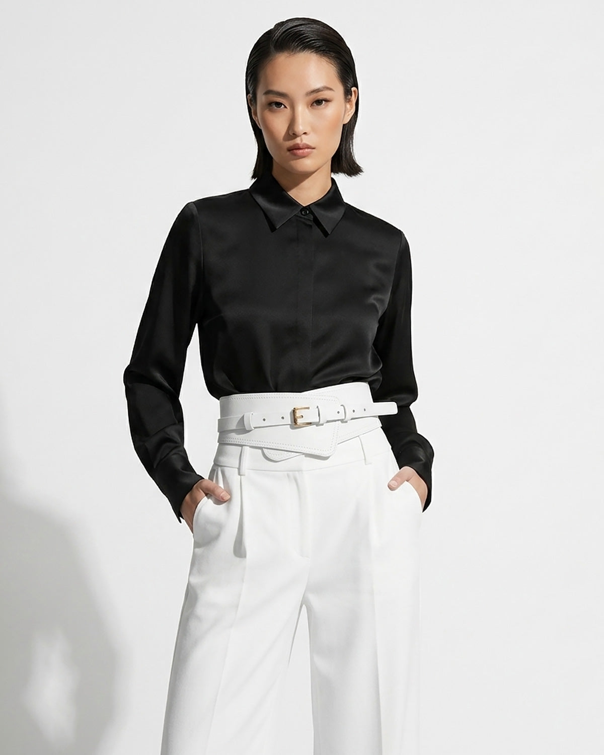 A model wearing a black satin long-sleeve button-down shirt tucked into high-waisted white trousers with a matching 2‑in‑1 White Women’s Leather Wide Transformable Statement Belt with Gold Buckle | BUCKLE Collective
