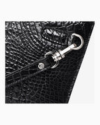 Close-up of a black crocodile leather clutch with a silver metal clasp and strap attachment on a light gray background | BUCKLE Collective