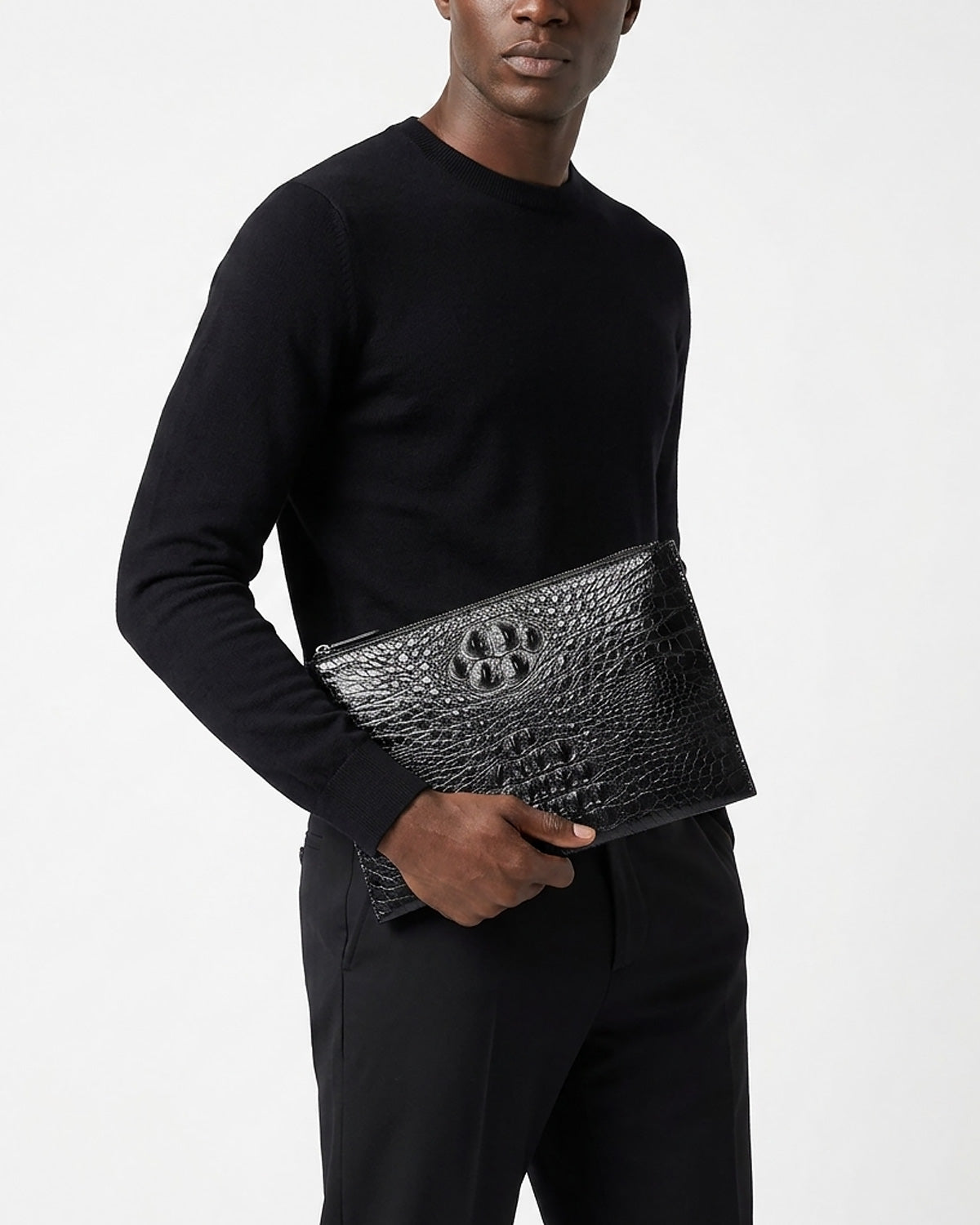 A man wearing a black long-sleeve crewneck sweater and black trousers holds a Black Genuine Siamese Crocodile Leather Envelope Clutch | BUCKLE Collective