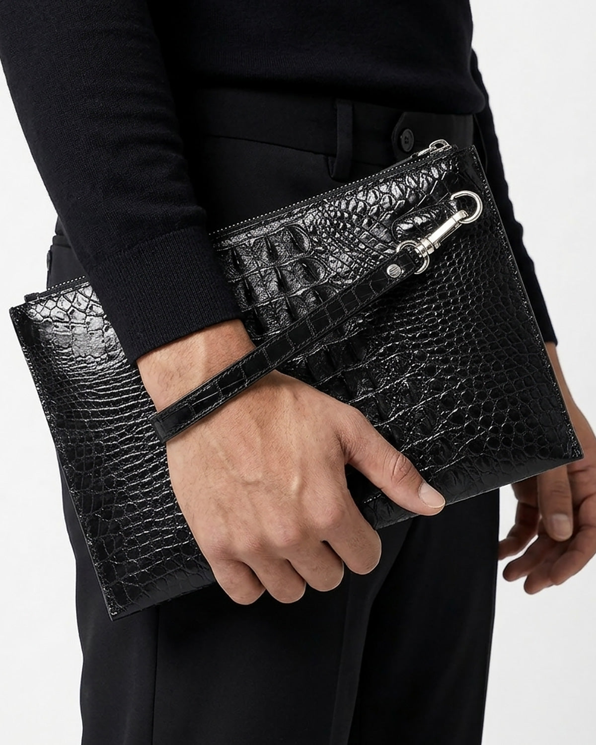A black genuine Siamese crocodile leather clutch bag with a matching wristlet strap and silver-tone hardware, held by a man wearing a black long-sleeve top and black trousers | BUCKLE Collective