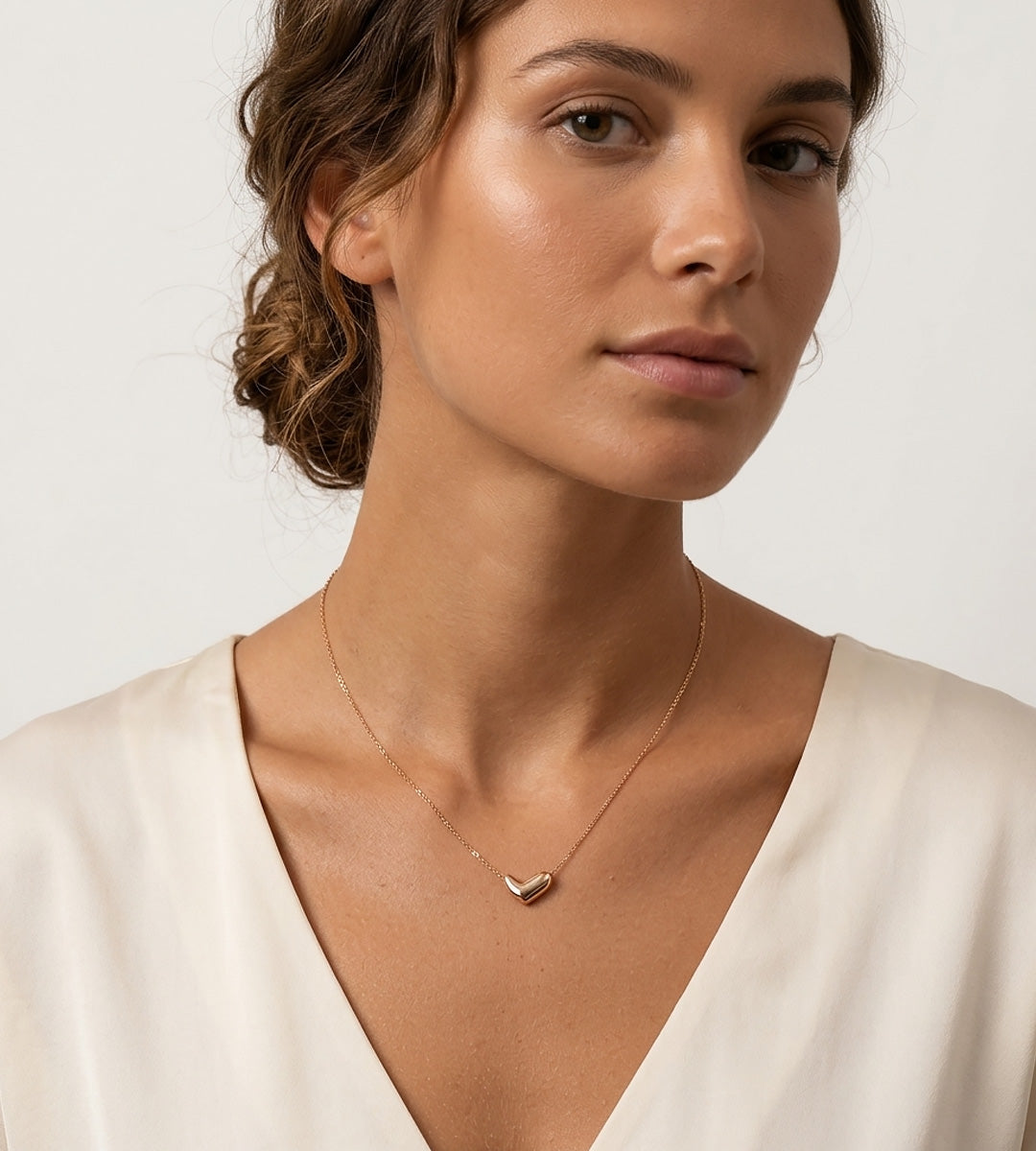 A close-up of a woman wearing a delicate 18K gold-plated S925 Silver Dainty Curved Bean Heart Necklace. She is wearing a cream-colored V-neck classic dress | BUCKLE Collective