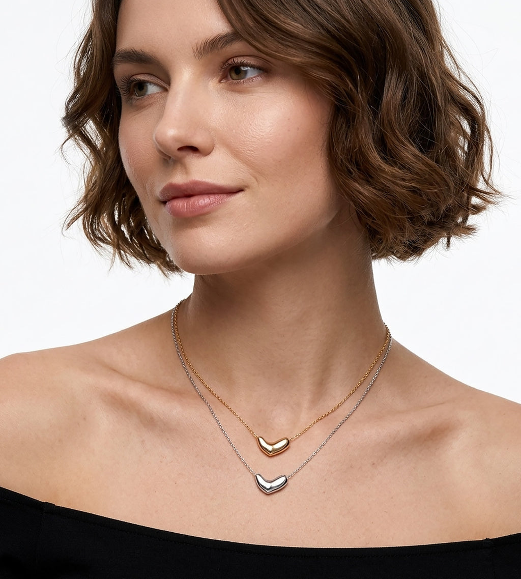 A close-up of a woman wearing two delicate necklaces: a gold chain with a small 18K gold-plated silver heart-shaped bean pendant and an 18K white-gold-plated silver chain with a small heart-shaped bean pendant | BUCKLE Collective
