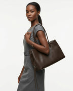 A woman wearing a gray dress carries a large dark brown leather commuter tote bag over her shoulder. BUCKLE Collective