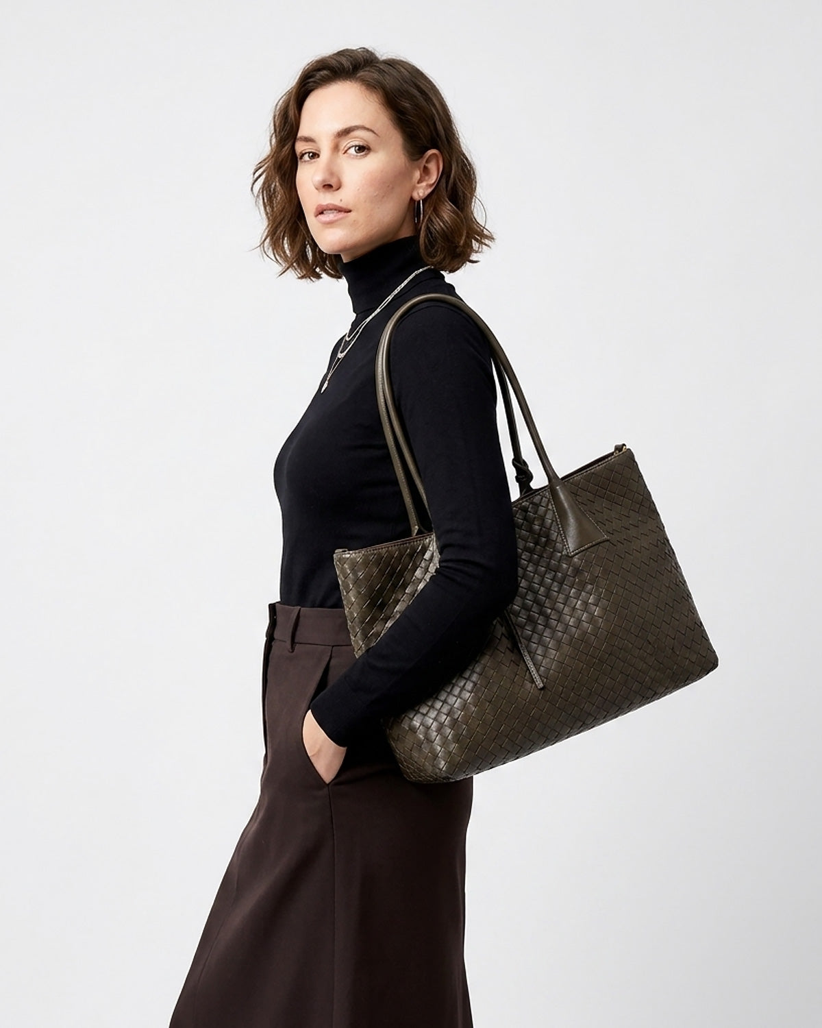 A woman wearing a black turtleneck and dark brown skirt carries a large woven dark green tote bag over her shoulder